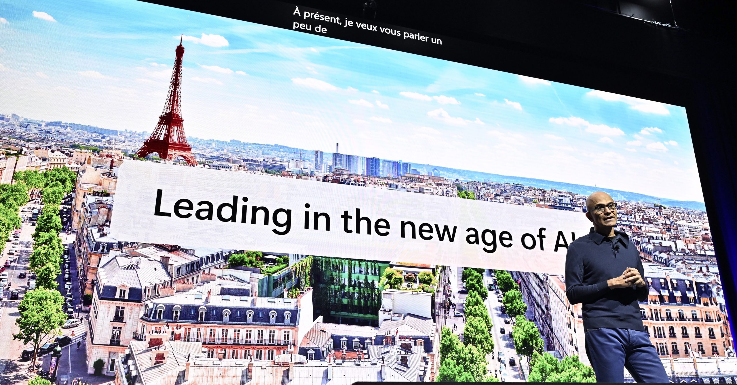 Satya Nadella, wearing a blue shirt standing in front of a screen with Eiffel Tower in frame and text that says Leading in the new age of AI