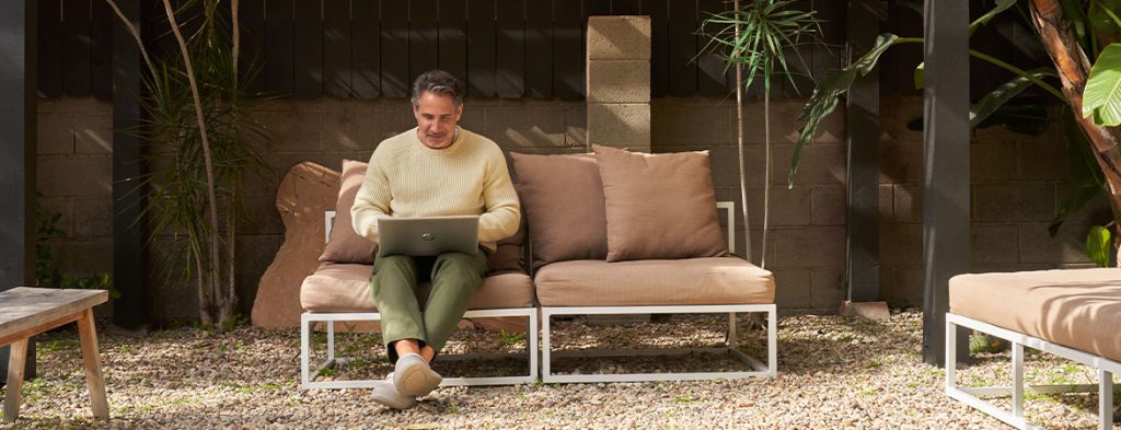 A man sits on a couch holding a laptop with plants in the background