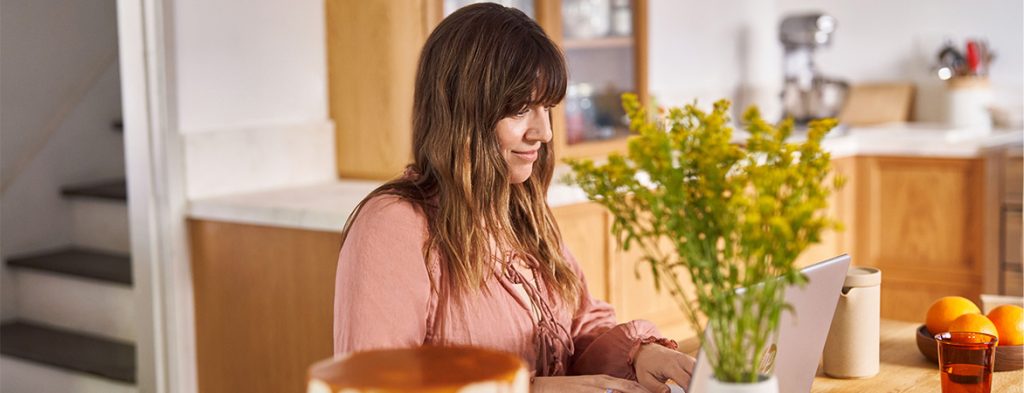 A woman sits at a table near a kitchen working on a laptop