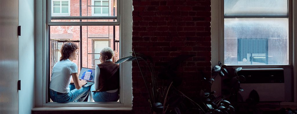 Two women sit in a windowsill around a laptop