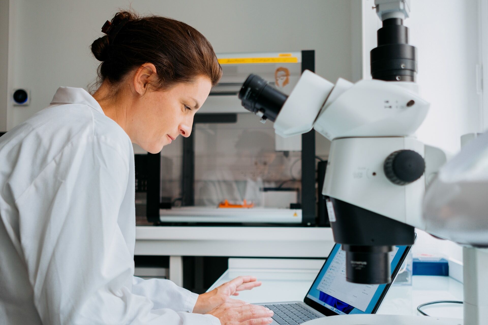 A woman with tied-up hair, wearing a white lab coat, works on a laptop in a laboratory, with a microscope in the foreground.