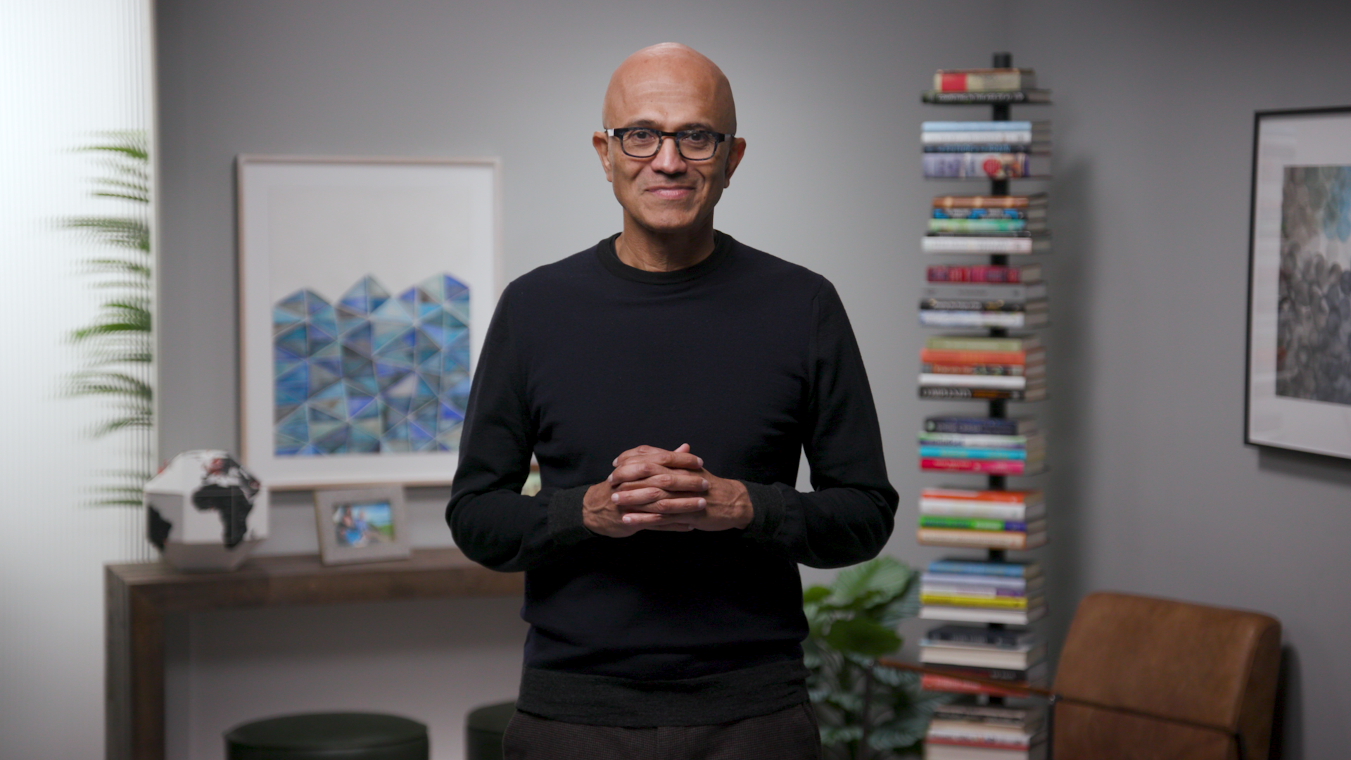 A man (Satya Nadella) in a black full sleeves shirt with his hands joined. Background shows a bookshelf and a picture