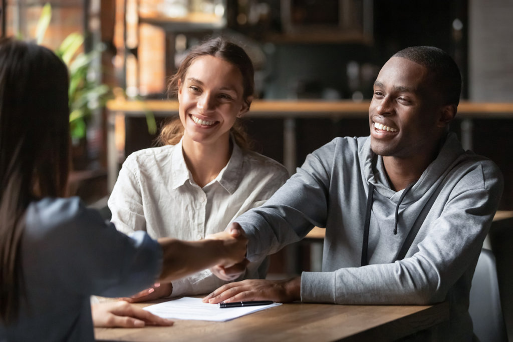 Happy young couple shake hand of bank manager broker buy insurance services take mortgage loan, mixed ethnicity customers handshake agent make agreement financial business deal at meeting