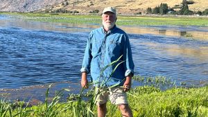 Bearded man in blue shirt in front of a lake and a strip of green marsh plants