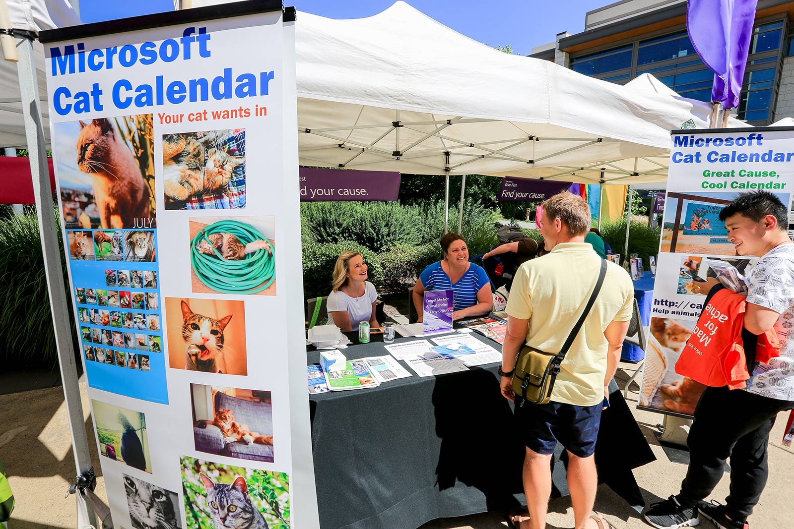 Two women sitting at an outdoor booth with large posters of cat pictures and title Microsoft Cat Calendar on either side